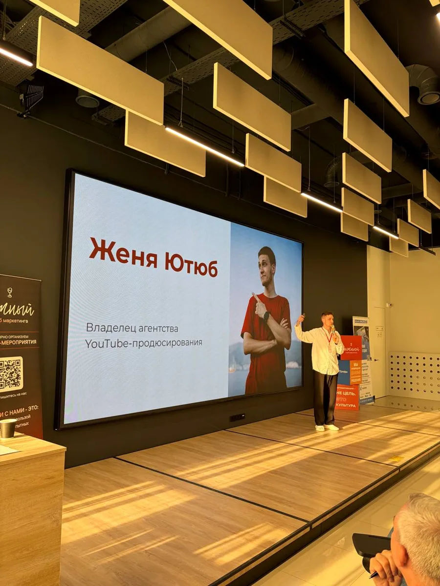 Evgeny Maximov on stage at a marketing club event, with his name and title as a YouTube production agency owner on the screen behind him