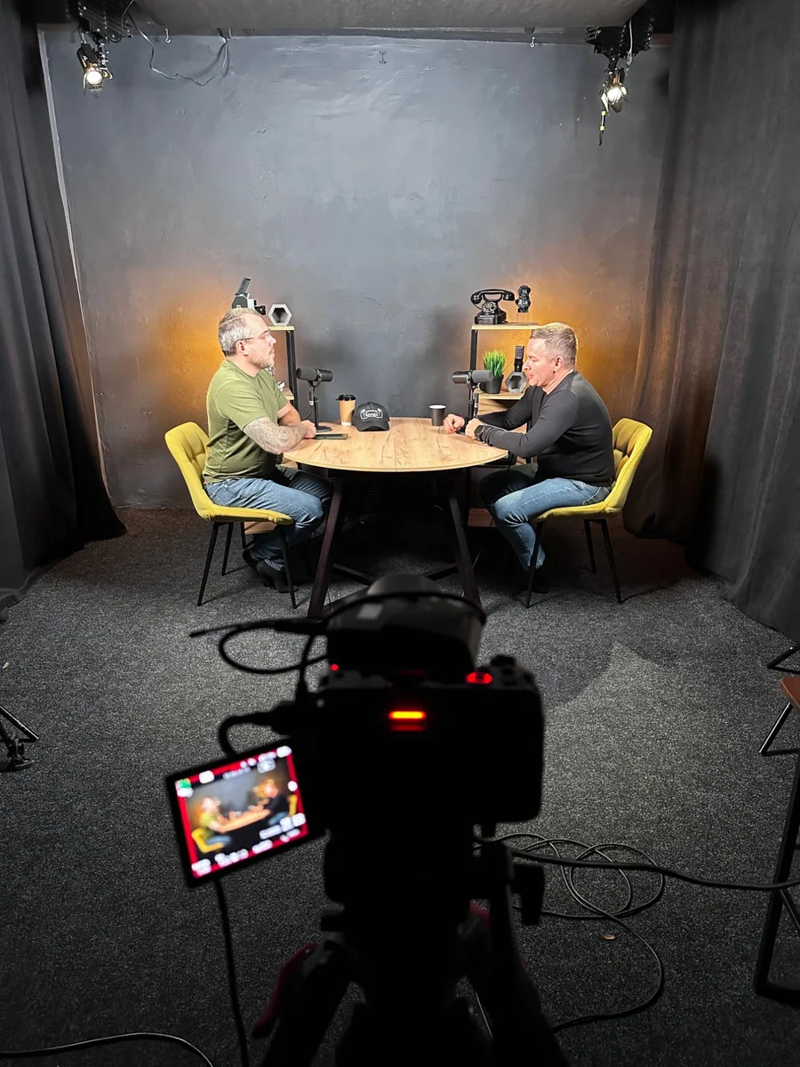 A podcast recording in a dark studio: two men at a round wooden table with microphones and yellow chairs, lit by spotlights, a camera on a tripod in the foreground