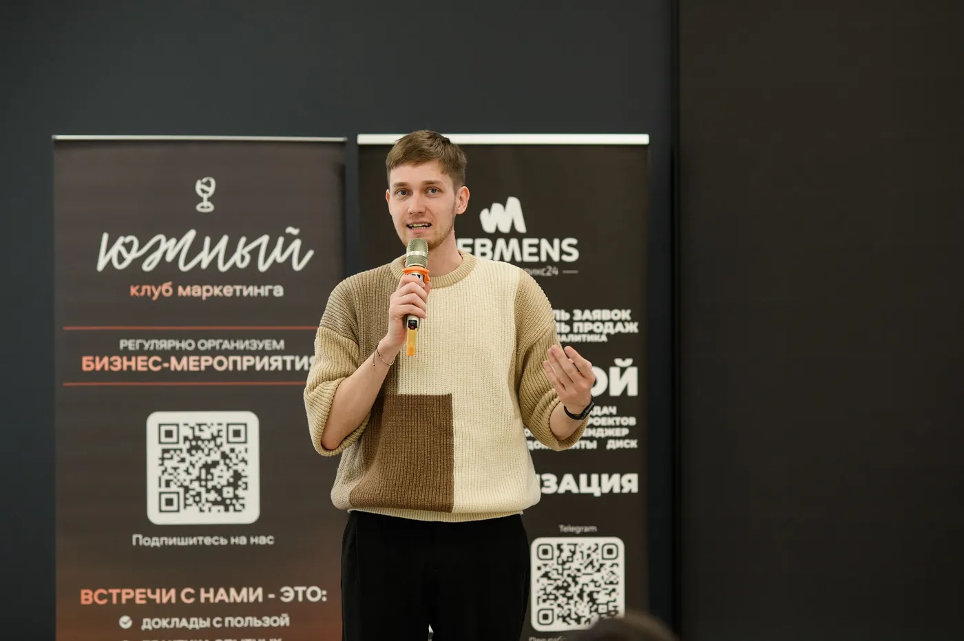 Evgeny Maximov speaking with a microphone at a marketing club event in Krasnodar, event banners behind him