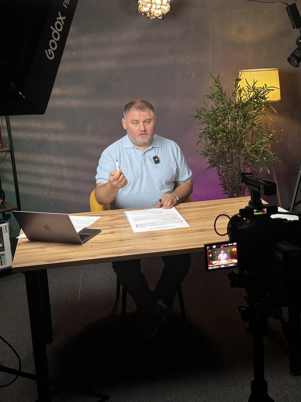 A client on set at a desk with a printed script in front of him, a camera and a softbox light around