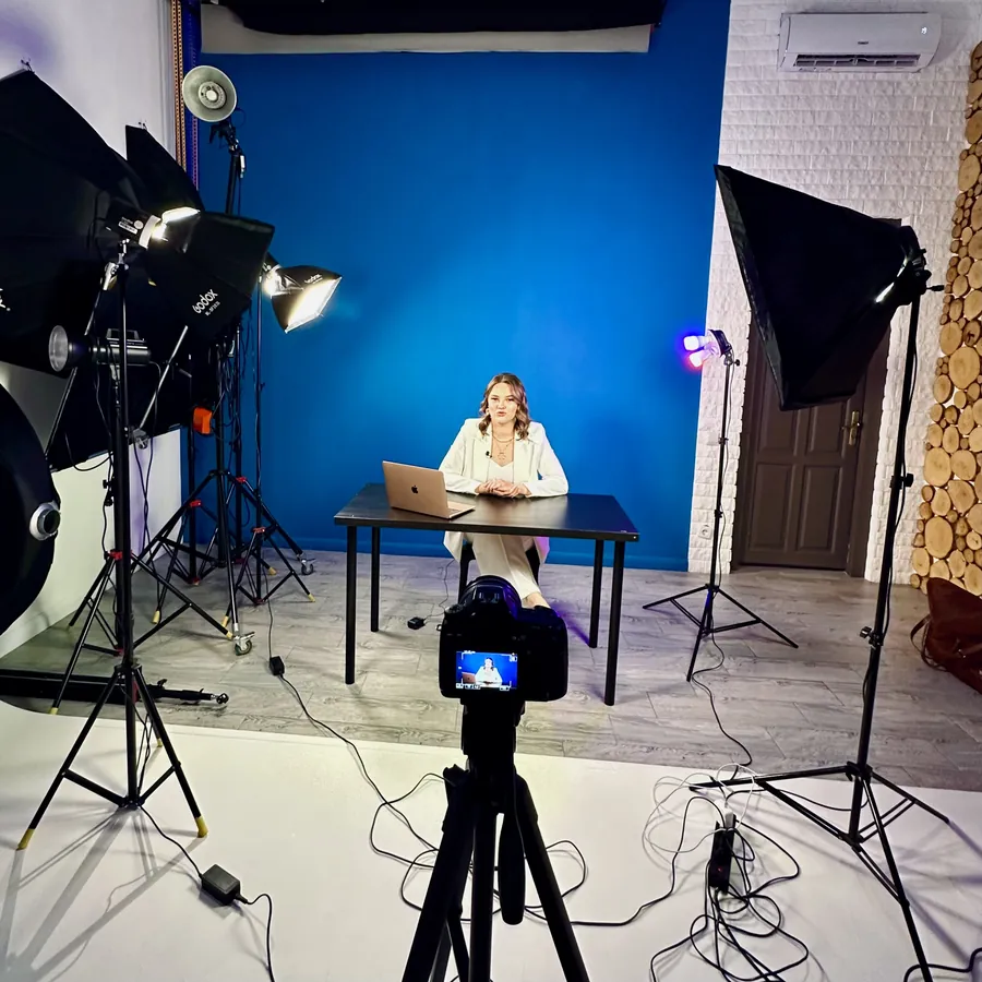 A studio shoot with a blue backdrop, softbox lights and a camera on a tripod in front of a client at a desk